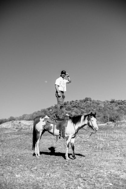 Man standing in a horse saddle wearing Mallard & Friends Black Five Panel Hat