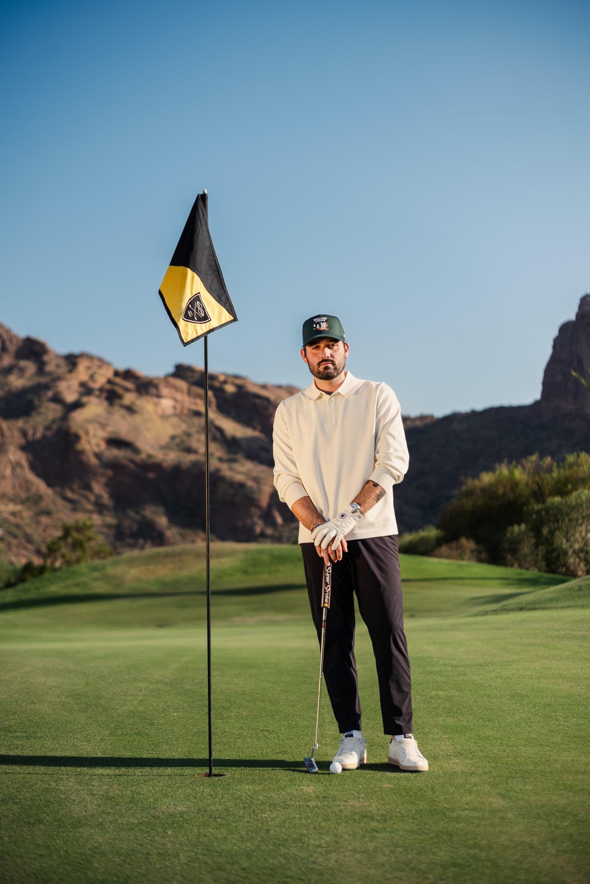 Man posing next to a golf hole flag wearing unique and stylish Mallard & Friends Dark Green Five Panel Hat