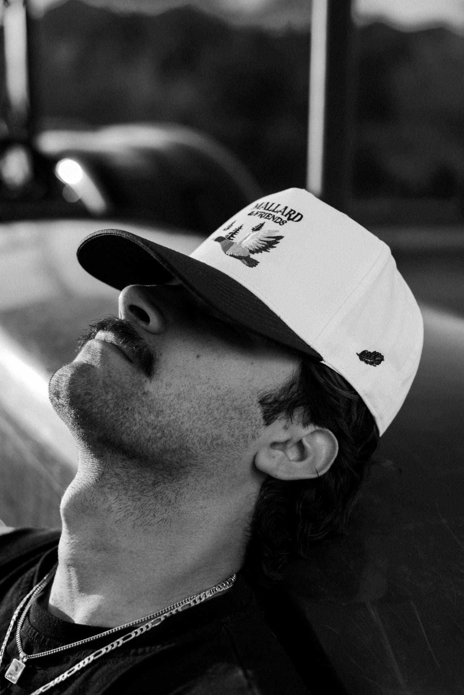 Black and white photo of a relaxed man in a tractor wearing Mallard and Friends white and Green Five Panel Hat 