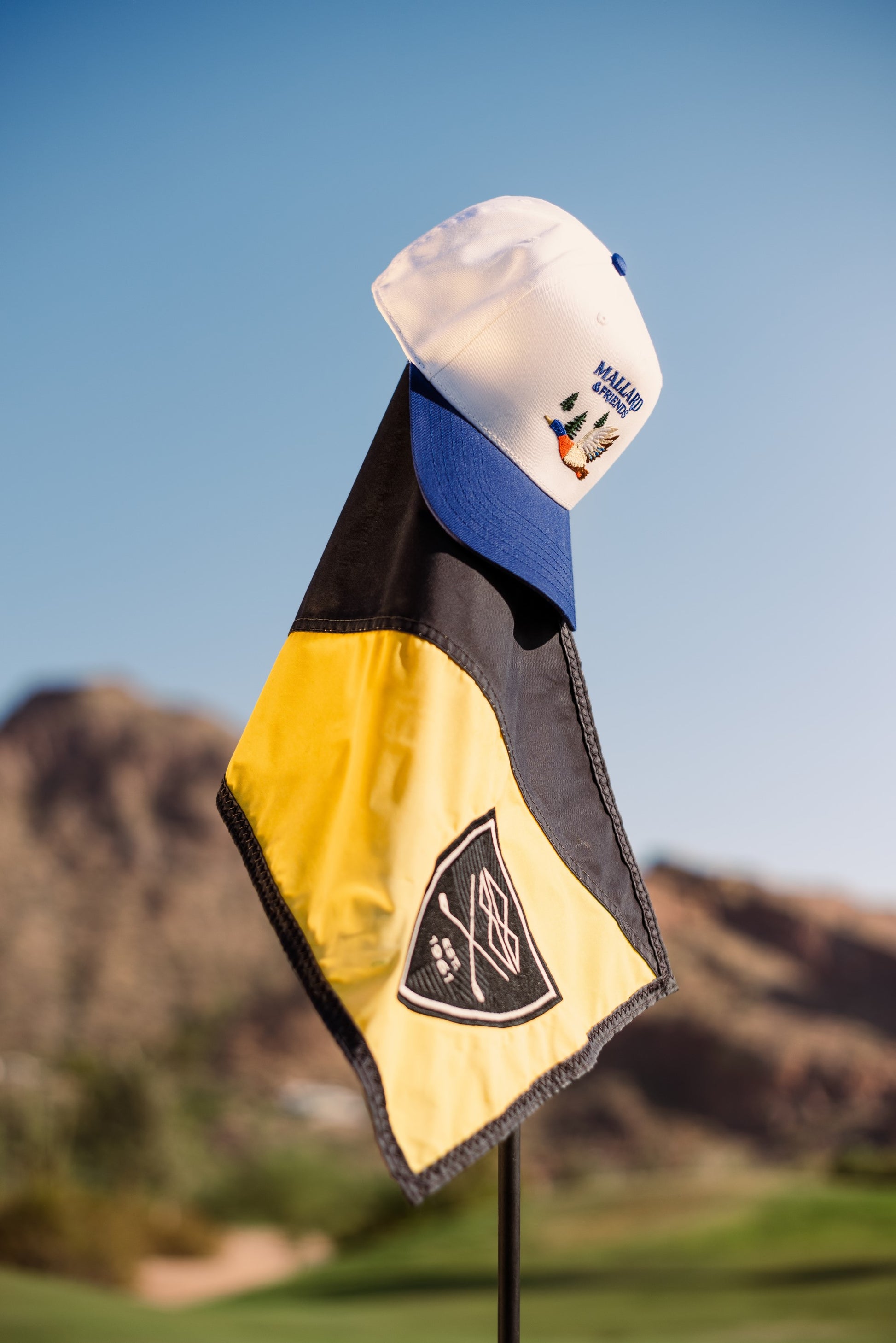 Mallard and Friends White and Royal Blue Five Panel Hat Hanging from the Golf Hole Flag